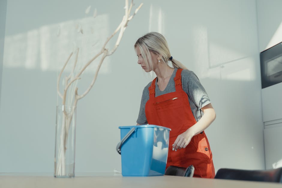 A professional cleaner from Cleaner Knightsbridge wearing a grey t-shirt and red apron, standing in a well-lit room with white and light grey walls, preparing for surface cleaning. The cleaner appears focused, with a blue cleaning bucket in front of her, likely containing cleaning supplies. The room features a ceramic tiled or laminated surface on the table, and in the background, there is a modern white appliance or storage unit. The space is bright, with natural light casting soft shadows, emphasizing the cleanliness and order of the environment, consistent with deep cleaning and sanitisation practices for residential and commercial spaces in Knightsbridge.