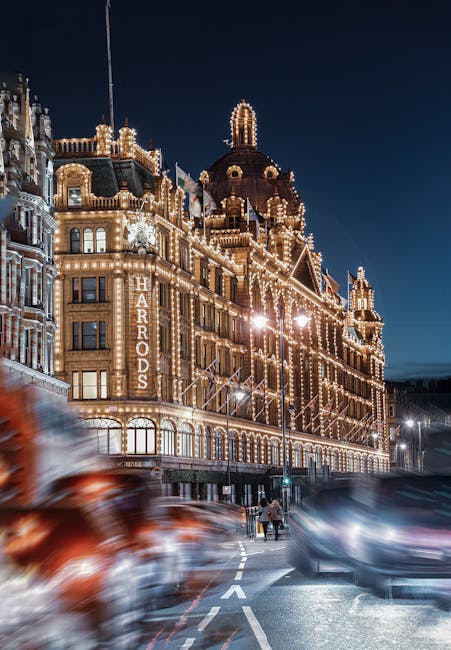 A nighttime view of the Harrods department store on Brompton Road in Knightsbridge, illuminated with warm white decorative lights outlining its historic architecture. The building's ornate facade features large windows, intricate detailing, and a central dome topped with a decorative spire. In the foreground, blurred moving cars indicate busy traffic, while pedestrians walk along the sidewalk under streetlights. The contrast between the dark evening sky and the bright, lit-up building highlights its grandeur, emphasizing the luxurious retail environment typical of Knightsbridge. This image showcases the store's elegant exterior, related to cleaning and maintenance services such as those offered by Cleaner Knightsbridge for commercial properties.