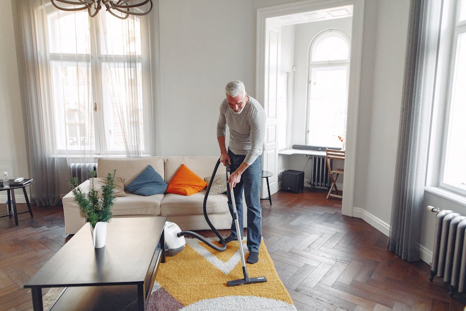 An older man with gray hair and glasses is engaged in surface cleaning within a well-lit living room that features wooden flooring laid in a herringbone pattern. He is vacuumming a yellow area rug with a modern design, using a black vacuum cleaner with a long hose. The room is furnished with a white sofa adorned with colorful cushions in blue and orange, positioned against large windows with sheer and heavier curtains, allowing natural light to fill the space. A small side table with a plant, a coffee table, and a desk near the window are visible, along with a radiator and some electronic equipment. The clean and organized room showcases a professional approach to domestic cleaning performed by [COMPANY_NAME], highlighting the importance of thorough surface cleaning and maintaining hygiene in a residential setting, consistent with the context of Brompton Road carpet cleaning cost and tips.