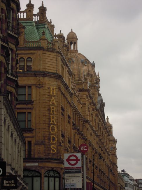 Exterior view of a historic sandstone building in Knightsbridge, featuring ornate architectural details and a green rooftop. The building's façade displays the sign 'HARRODS' in tall yellow lettering. In the foreground, a London Underground station sign and bus stop icons are visible. The scene is under cloudy sky lighting, with part of the street visible. This image is related to cleaning and maintenance of commercial venues, with Cleaner Knightsbridge specializing in deep cleaning services for such iconic locations.