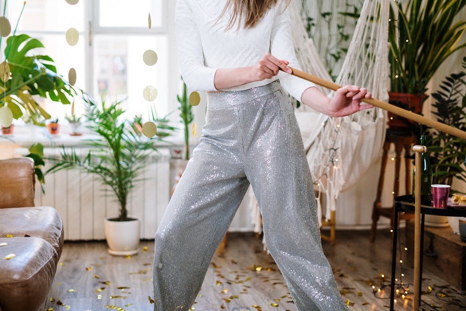 A person wearing silver sequin trousers and a white top is sweeping a living room with a long wooden broom handle. The room features a wooden floor scattered with gold confetti, a brown leather sofa on the left, and several potted plants including a large green one near a window and a smaller one on a side table. In the background, white walls are decorated with hanging ornaments and fairy lights, and a peach-colored disposable cup is placed on a side table. The space appears bright and tidy, and the scene depicts domestic deep cleaning to maintain hygiene after a celebration, in line with services provided by Cleaner Knightsbridge.