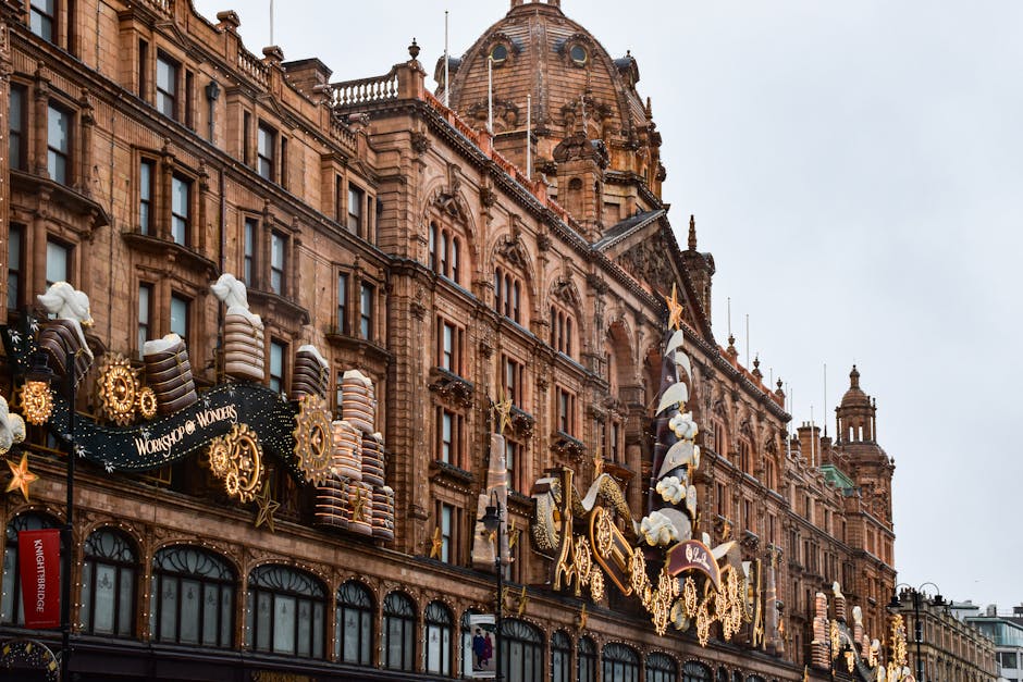 Exterior view of Harrods department store in Knightsbridge, featuring ornate architectural details made of red brick with decorative stone elements. The building is decorated for the festive season with large, illuminated ornaments, stars, and themed displays on the upper facade, including large gingerbread house decorations and candy cane motifs. The windows extend across several floors, with a combination of arched and rectangular shapes, and the scene is captured during daytime with a slightly overcast sky. The overall appearance is clean and well-maintained, reflecting the luxury and grandeur associated with Harrods, but no cleaning tools or surfaces are visible in the image.