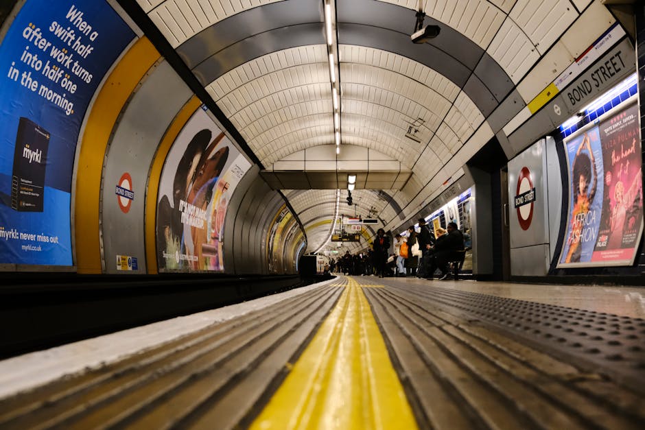 A low-angle view of Knightsbridge station platform showing the textured floor with a yellow safety line in the center. The platform is illuminated by fluorescent lighting embedded in the arched ceiling. The walls are lined with advertisements and station signage, including signals and information boards. Passengers are seated and standing near the platform edge, waiting for the train. The scene is clean, with no visible litter or debris, and the surfaces appear well-maintained and hygienic, consistent with professional cleaning standards provided by Cleaner Knightsbridge specializing in surface cleaning and sanitisation for busy transit environments.