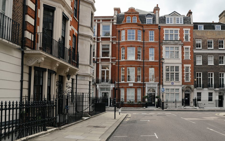 A street scene in Knightsbridge featuring a row of elegant, multi-story residential buildings with traditional brick and stone facades. The buildings have large sash windows, wrought iron balconies, and decorative architectural details. The pavement is clean with well-maintained black metal railings bordering the properties. The street includes a smooth asphalt road with marked parking spaces and a few street signs. Soft natural lighting highlights the pristine condition of the exteriors, suggesting regular surface cleaning and maintenance by Cleaner Knightsbridge as part of their deep cleaning services for homes in the area.