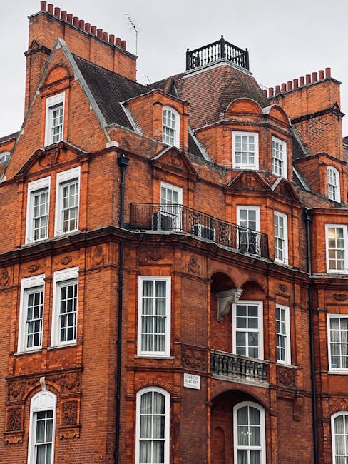 A street scene in Knightsbridge featuring a row of elegant, multi-story residential buildings with traditional brick and stone facades. The buildings have large sash windows, wrought iron balconies, and decorative architectural details. The pavement is clean with well-maintained black metal railings bordering the properties. The street includes a smooth asphalt road with marked parking spaces and a few street signs. Soft natural lighting highlights the pristine condition of the exteriors, suggesting regular surface cleaning and maintenance by Cleaner Knightsbridge as part of their deep cleaning services for homes in the area.