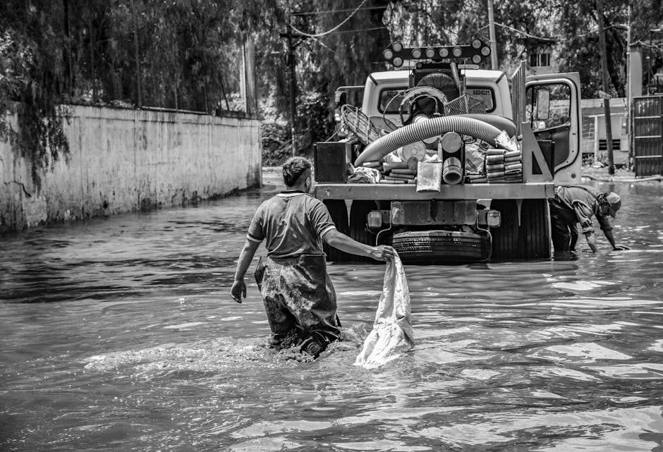 Two men are performing flood cleanup in an outdoor area, with one standing in water that reaches mid-calf level, holding a cloth or bag, and the other bending down near a large industrial water pump. 