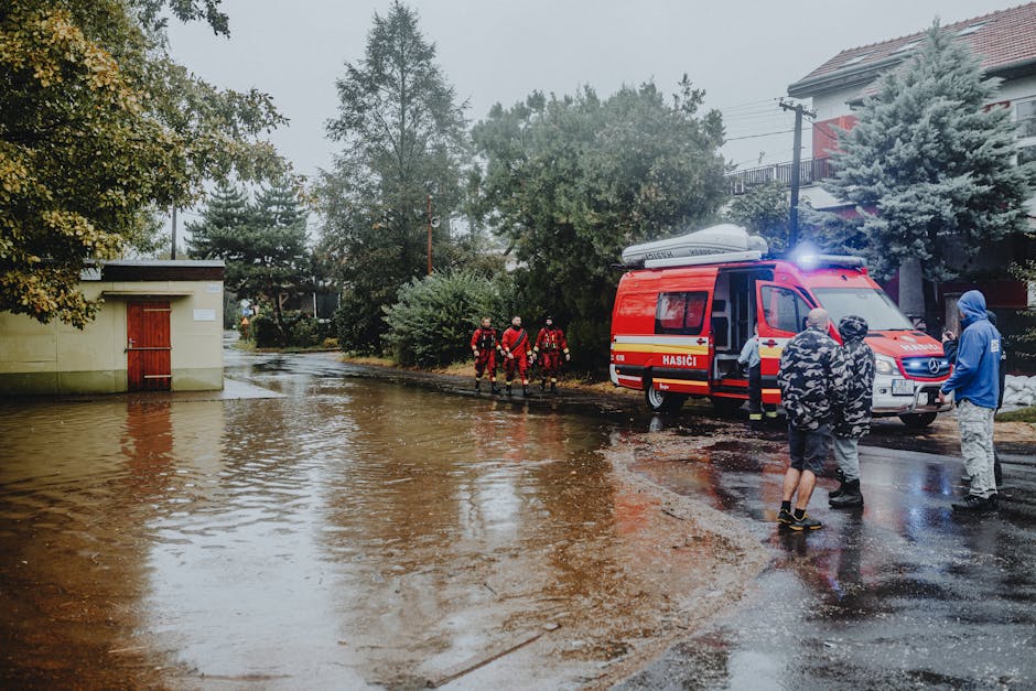 Two men are performing flood cleanup in an outdoor area, with one standing in water that reaches mid-calf level, holding a cloth or bag, and the other bending down near a large industrial water pump. 