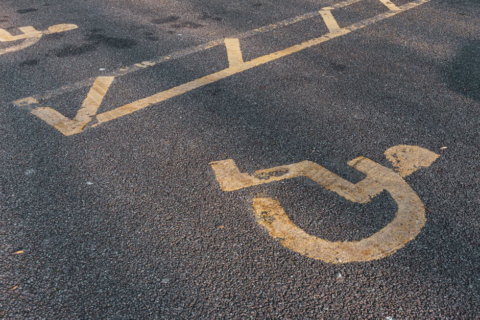 Close-up view of a parking space designated for disabled access on an asphalt surface, featuring a faded yellow wheelchair symbol painted on the ground. The area shows signs of wear and weathering, with visible cracks and rough texture on the surface. Surrounding the symbol, the asphalt is dark and slightly textured, with a few minor debris scattered around. The lighting is natural, highlighting the contrast between the paint and the asphalt. This image emphasizes the importance of designated parking areas for accessibility, and reflects the need for regular maintenance to keep markings visible. Cleaner Knightsbridge provides surface cleaning and maintenance services, ensuring these markings remain clear and accessible for all users, aligning with their focus on hygiene and surface upkeep for commercial and residential spaces. The photo is relevant to the 'What to know about access and parking for Knightsbridge cleaners' page on cleanerknightsbridge.co.uk, illustrating parking area features and the importance of proper surface care.